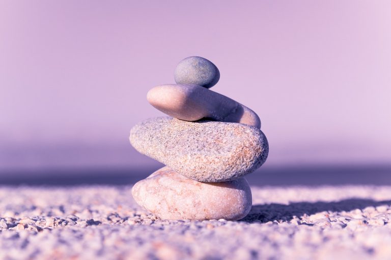 Four pebbles balancing on top of each other on a pebbled beach, against a light purple background