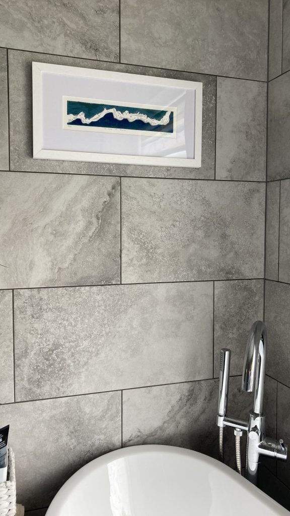 A Slice of Heaven lino reduction print in a grey frame hanging on a grey tiled wall above a white sink and tap