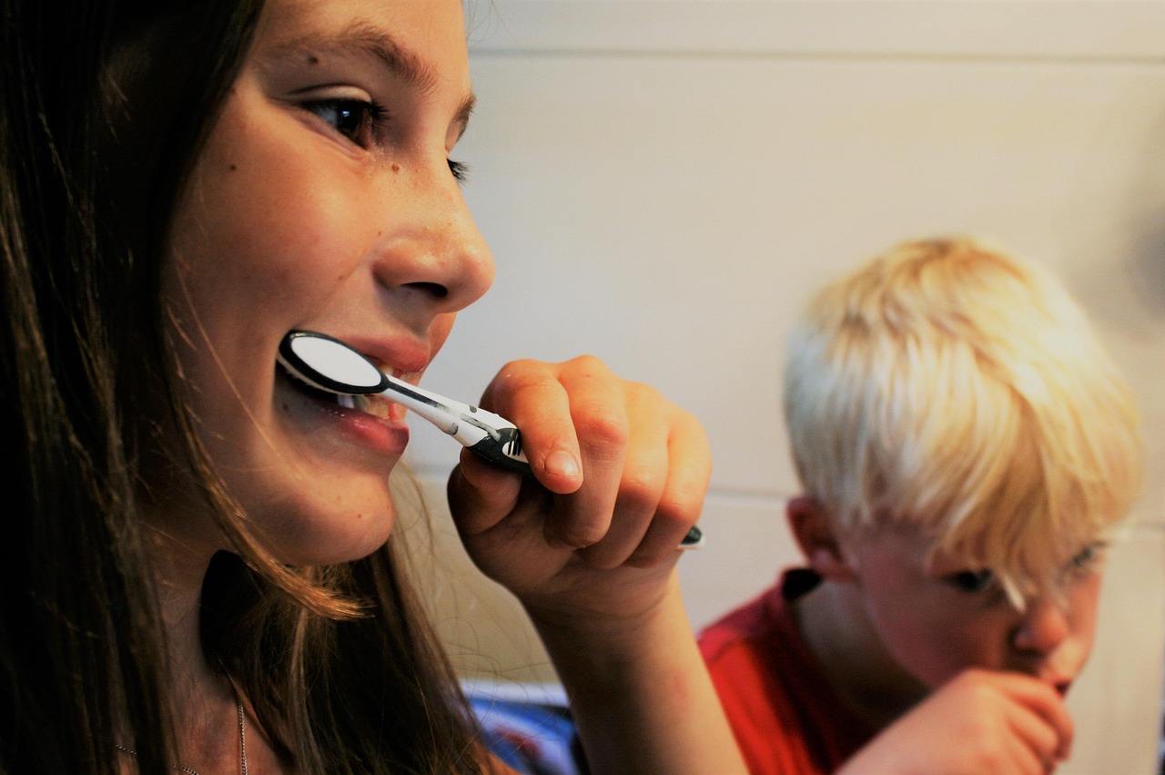 An older girl in the forefront of the shot, brushing her teeth, with a younger child brushing their teeth in the background