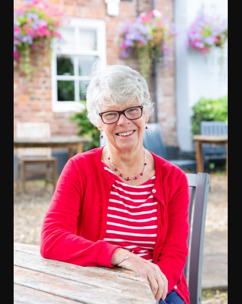 Headshot of Ann Girling wearing a red cardigan and red and white striped top, sitting at a wooden table, whose article shares tips for self-care for busy parents