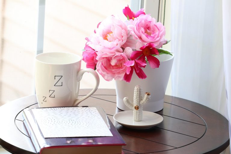 A small round wooden table with a white pot containing pink flowers, a notepad, a cup with ZZZ on and a small ceramic cactus ornament