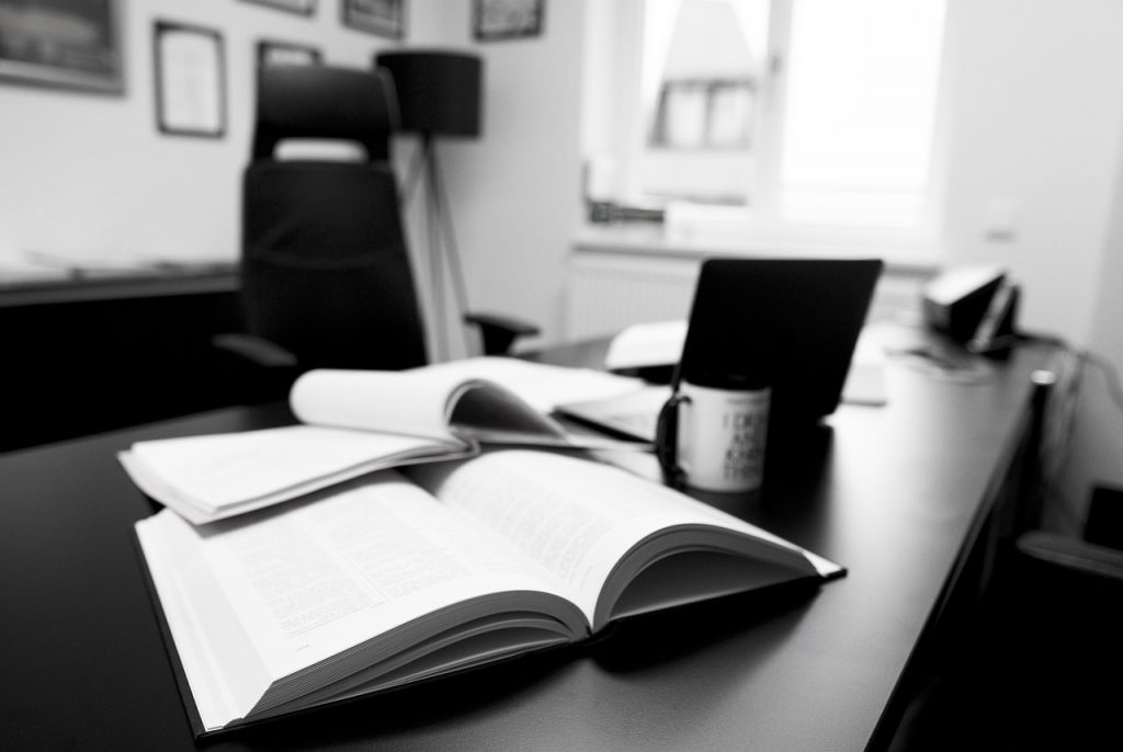 Black and white image of a desk with open text books, cup and open laptop. Window and chair in the background