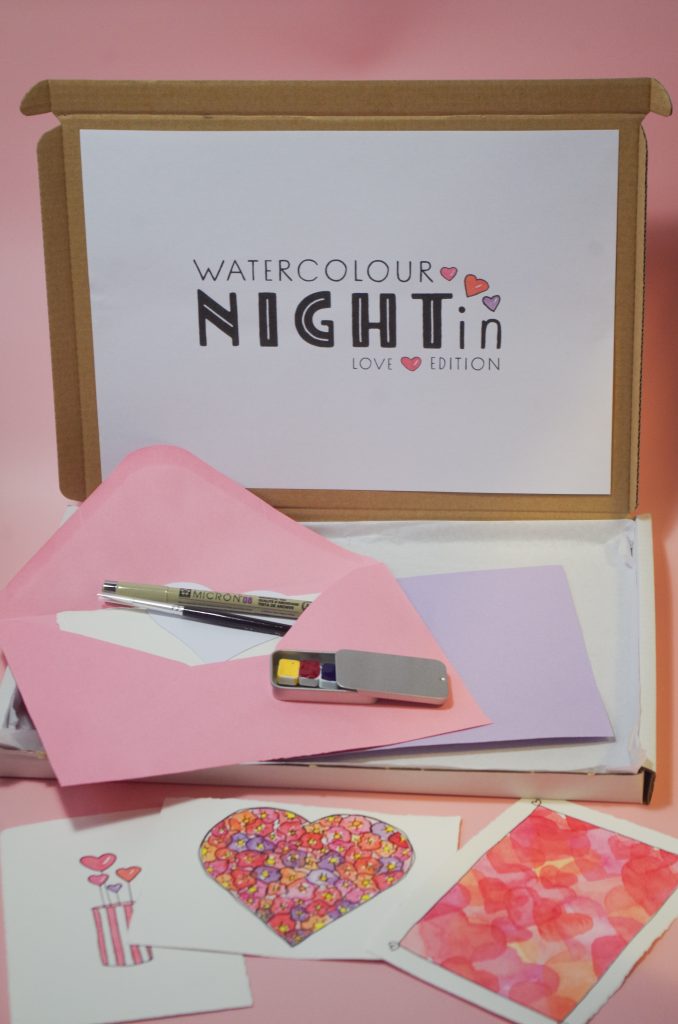 A box in the background with the words "Watercolour Night In" across the front, with paints, brushes and watercolour paintings in the foreground