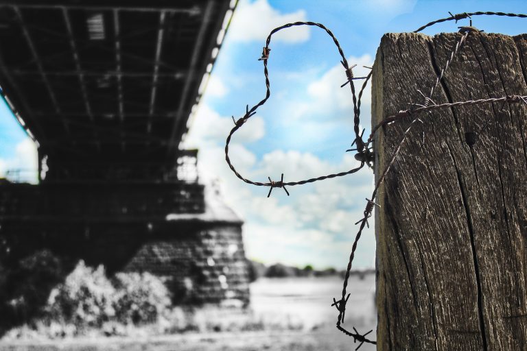 A heart shaped created from a piece of barbed wire in the forefront of the image with a bridge and scenery in the background