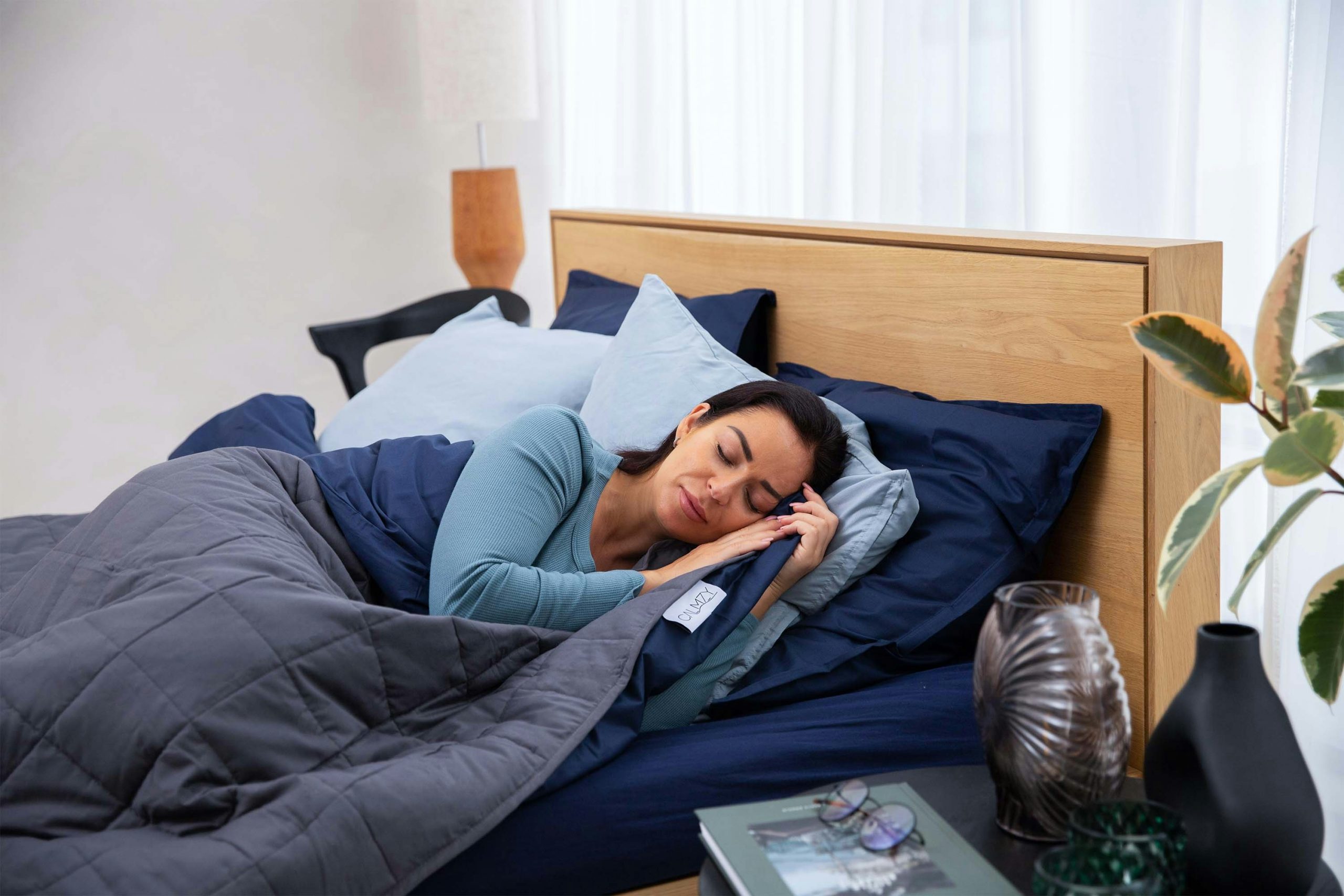A woman lying on her side asleep in a bed with blue pillows and cover, and white curtains behind the bed