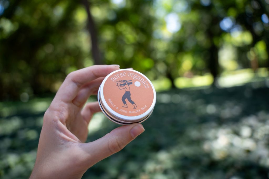 Womans hand holding a tine of Muscle & Joint Builder Balm with trees and a path in the background