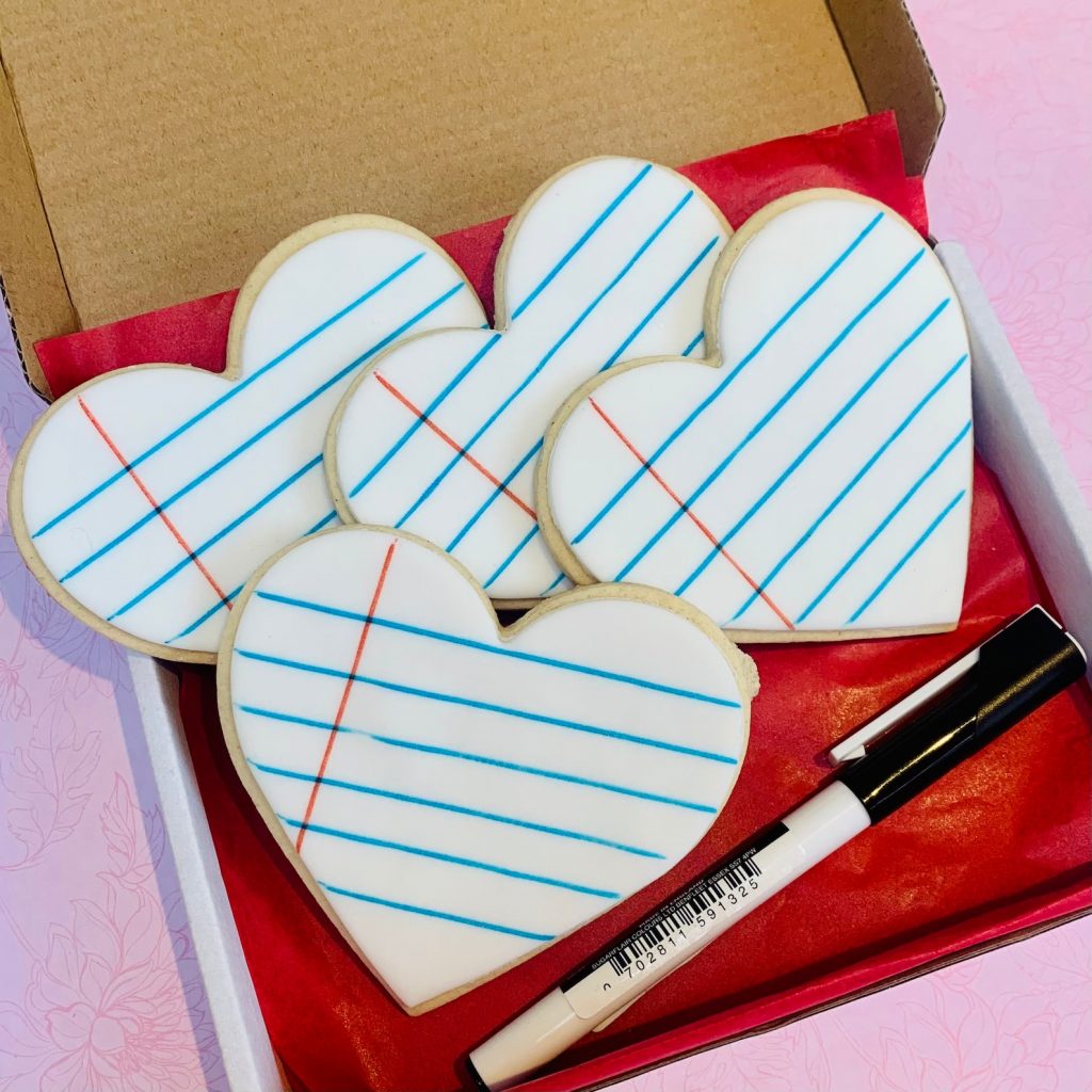 4 heart-shaped biscuits with white icing and lines on them, displayed in a box with a pen beside them
