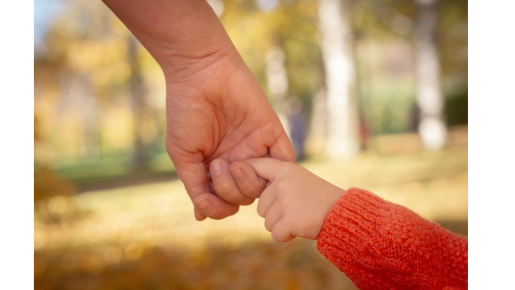 A man's hand holding a young child's finger against a background of trees